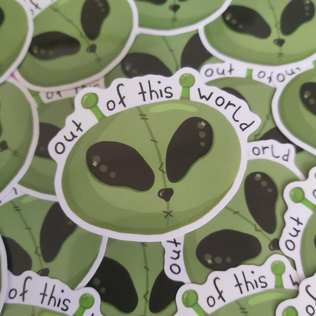 A close-up of a die-cut sticker featuring a cartoon green alien head with stitched details and the phrase 'out of this world' in black lettering, surrounded by a white border. Multiple identical stickers are layered in the background.