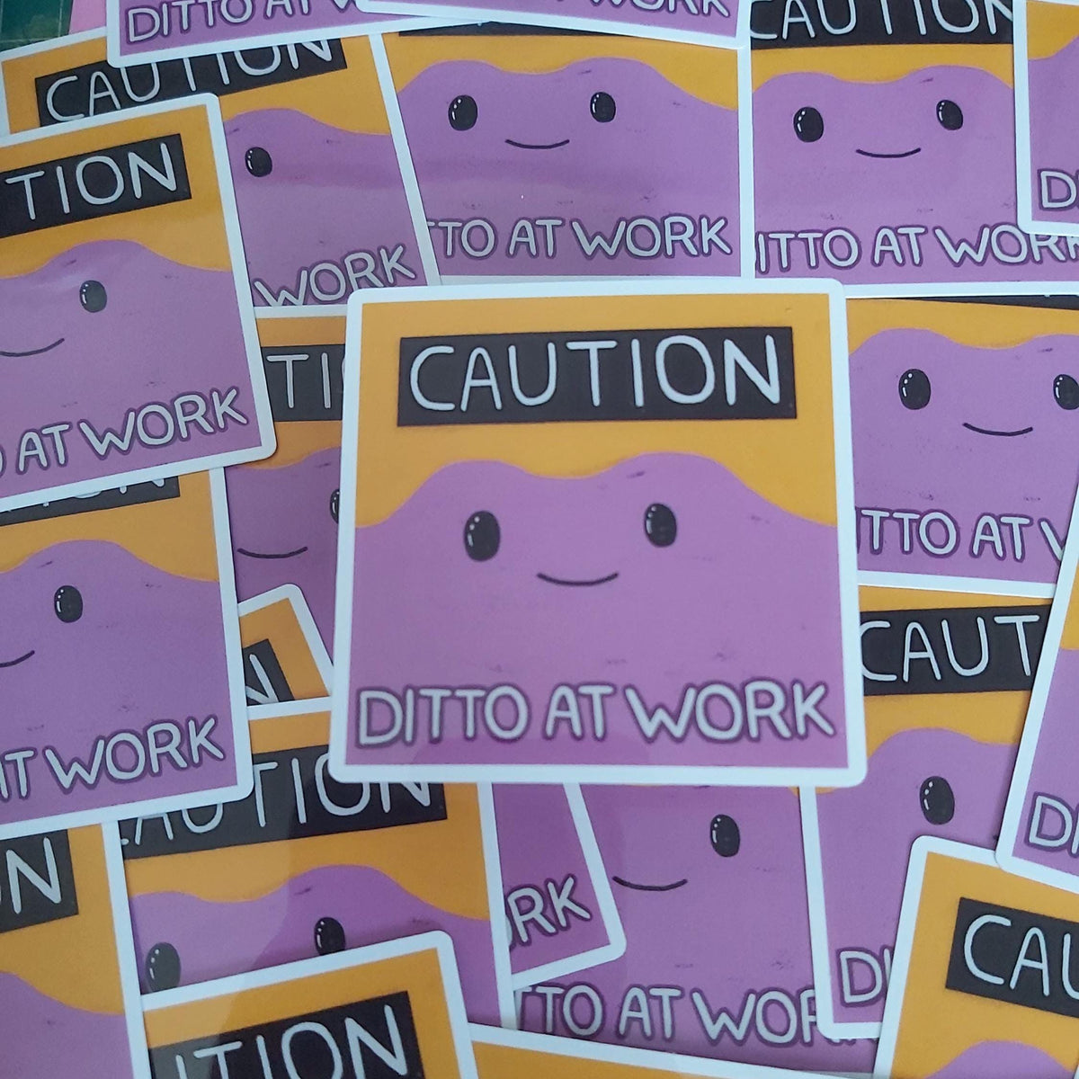 A pile of die-cut stickers featuring a cartoon purple blob face with black eyes and a smile, set against a yellow background with a black banner reading 'CAUTION DITTO AT WORK'.