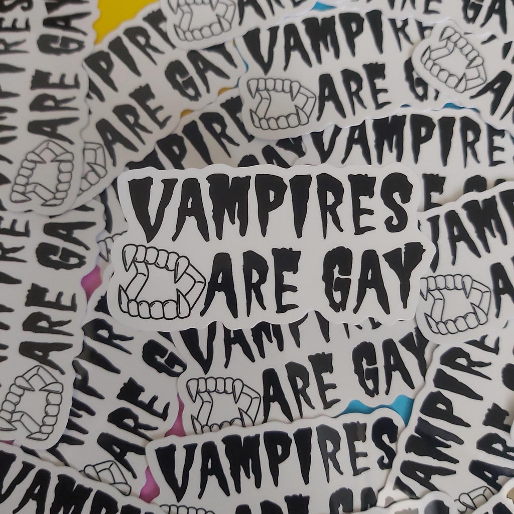 A pile of white die-cut stickers featuring the text 'VAMPIRES ARE GAY' in a black hand-drawn font, accompanied by black line drawings of vampire teeth. 