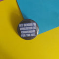 A round pinback button with the text 'MY GENDER IS WHATEVER IS CONVENIENT FOR THE BIT.' in white on a dark gray background, photographed on a colorful paper backdrop split between yellow, blue, and purple.