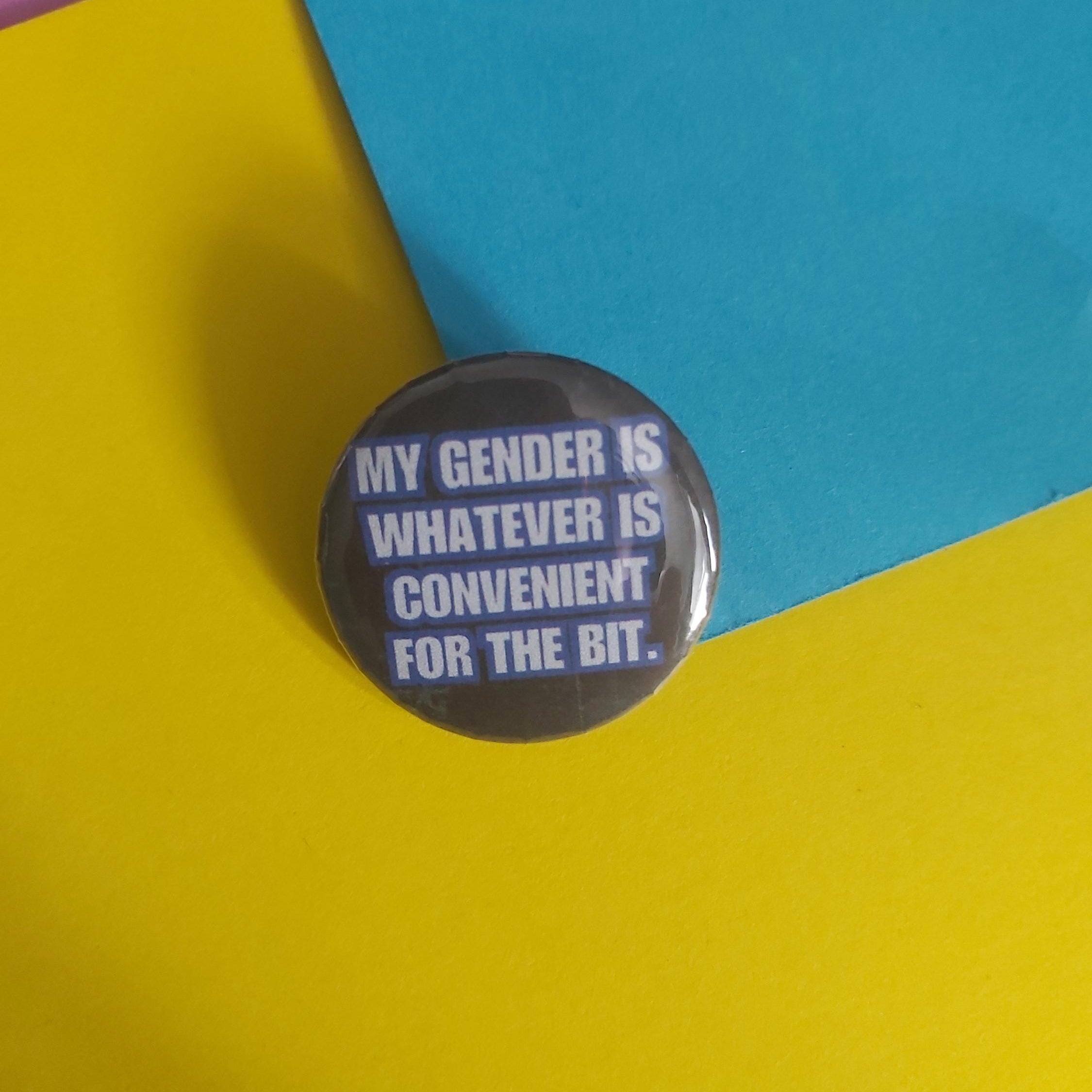 A round pinback button with the text 'MY GENDER IS WHATEVER IS CONVENIENT FOR THE BIT.' in white on a dark gray background, photographed on a colorful paper backdrop split between yellow, blue, and purple.