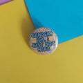 A round pinback button badge with the text 'GENDER? IDK, I JUST GOT HERE' in blue lettering over a purple line-drawn background, placed on yellow and blue paper.