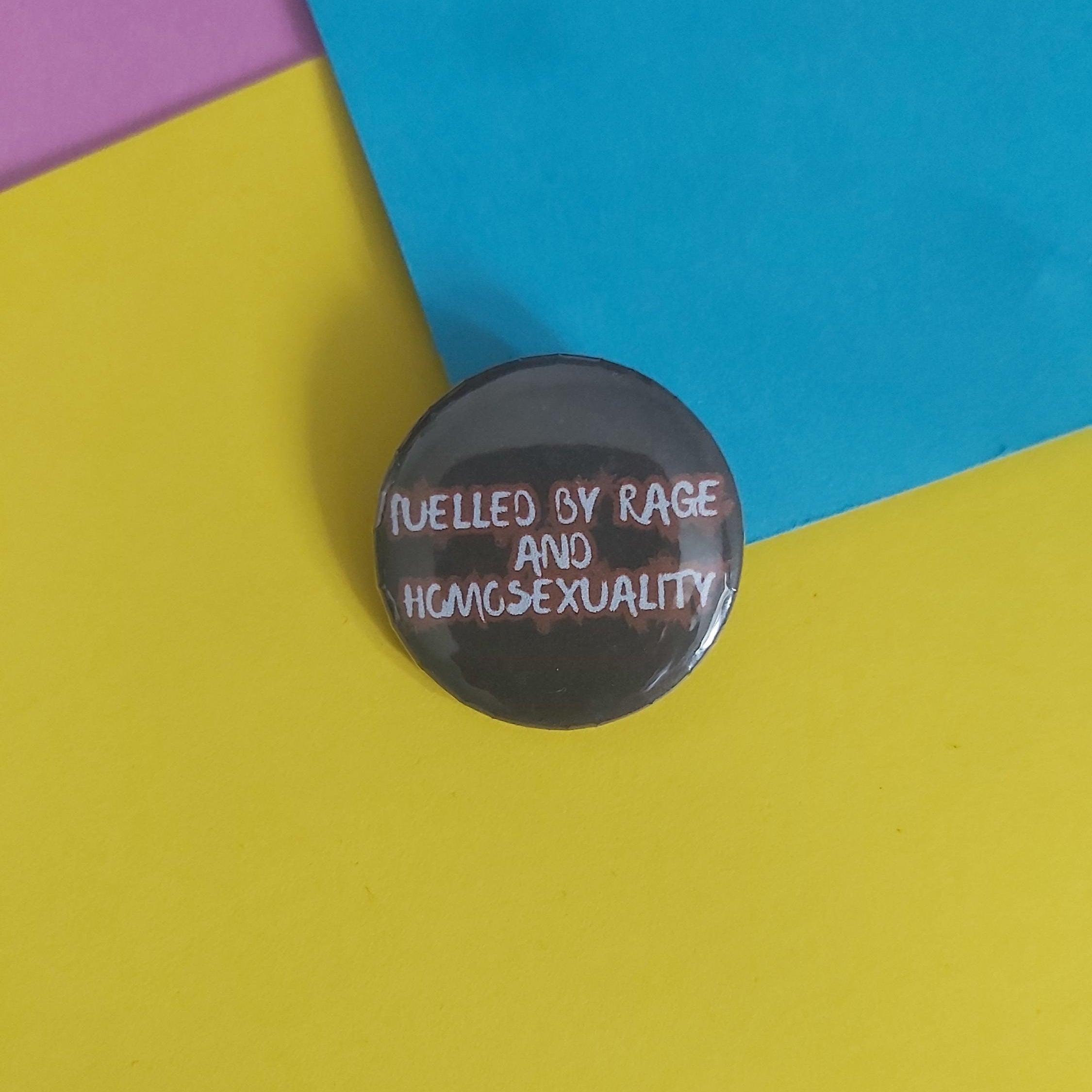 A round black pin badge with white text reading 'FUELLED BY RAGE AND HOMOSEXUALITY' placed on a colorful background of yellow, blue, and purple paper.