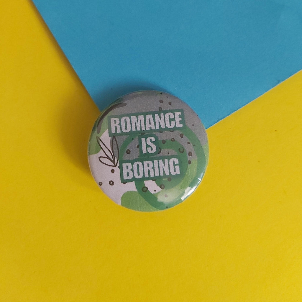 A round pin-back button badge with the text 'ROMANCE IS BORING' in white on a green and grey abstract background, placed on a yellow and blue paper backdrop.