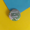 A round pin-back button badge with the text 'ROMANCE IS BORING' in white on a green and grey abstract background, placed on a yellow and blue paper backdrop.
