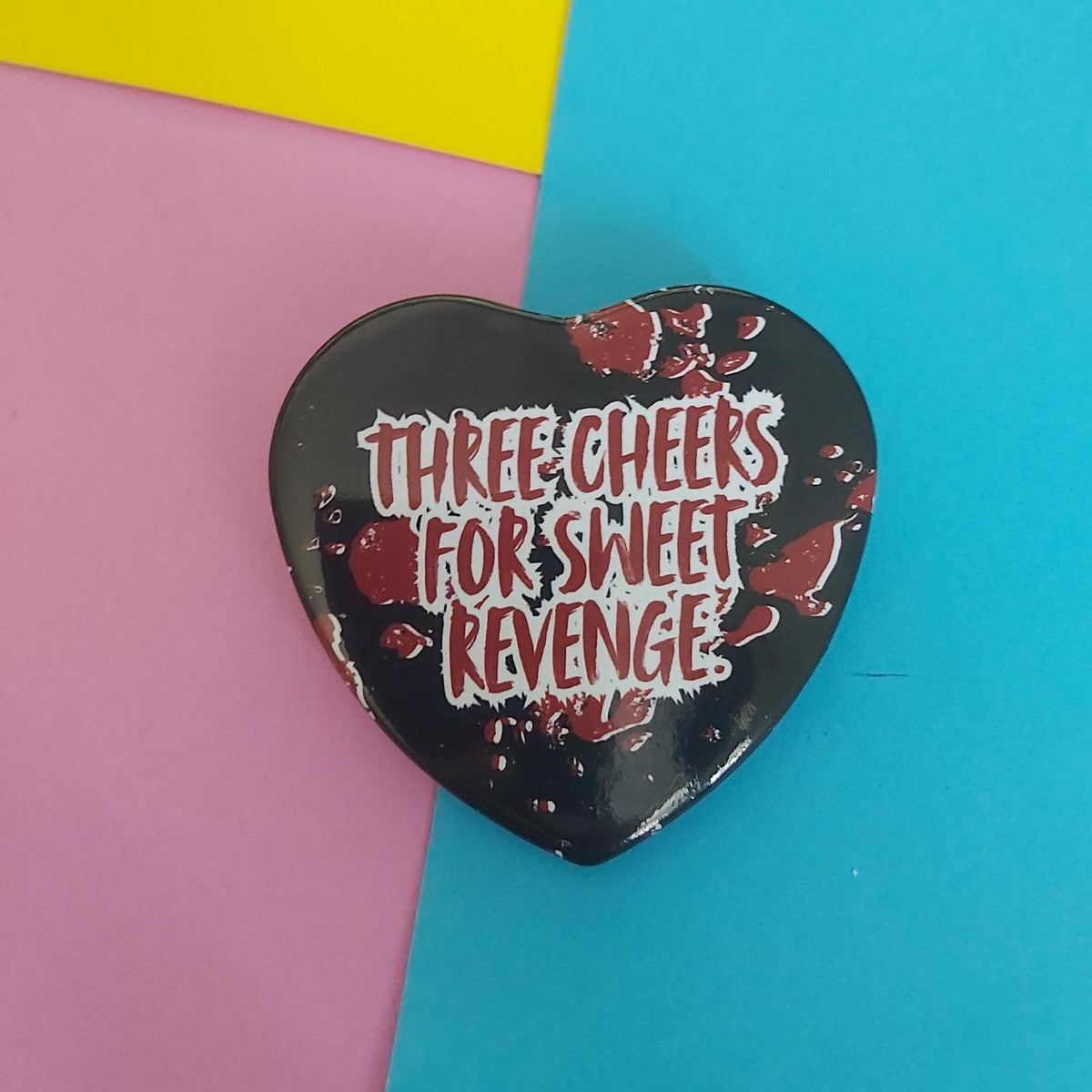 A heart-shaped black with the text 'THREE CHEERS FOR SWEET REVENGE' in white and red, styled with red splatter details, set against a colourful background of pink, yellow, and blue paper.