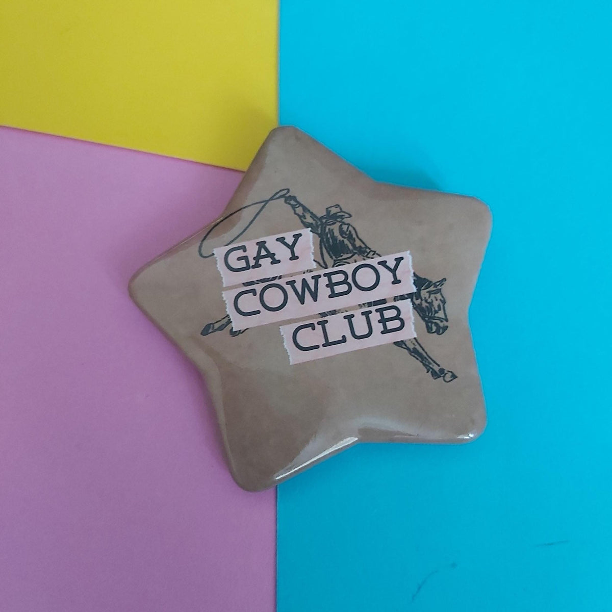 A star-shaped pin badge with a tan background, featuring a black cowboy illustration and the text 'GAY COWBOY CLUB' in white lettering, set against a background split diagonally into yellow, pink, and blue sections.