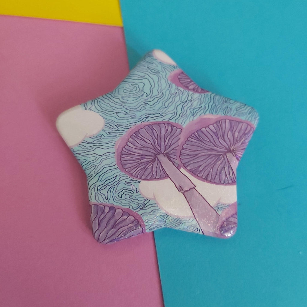 A star-shaped pin badge featuring a purple mushroom pattern over a light blue and white wavy background, displayed on a split pink and teal paper backdrop.