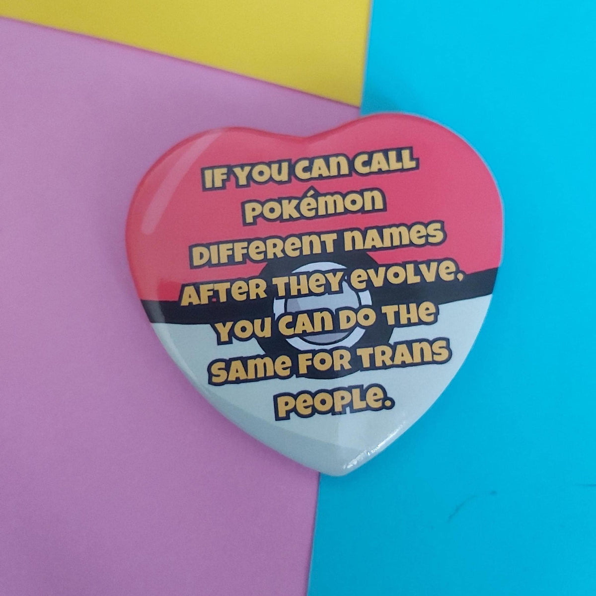 A heart-shaped Pokémon pin badge with a red, black, and white design and a transgender awareness message, displayed on a split pastel pink and blue background.