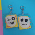 A reversible acrylic keychain with cartoon faces, one smiling and one sad, attached to silver keyrings, displayed on a bright blue background.