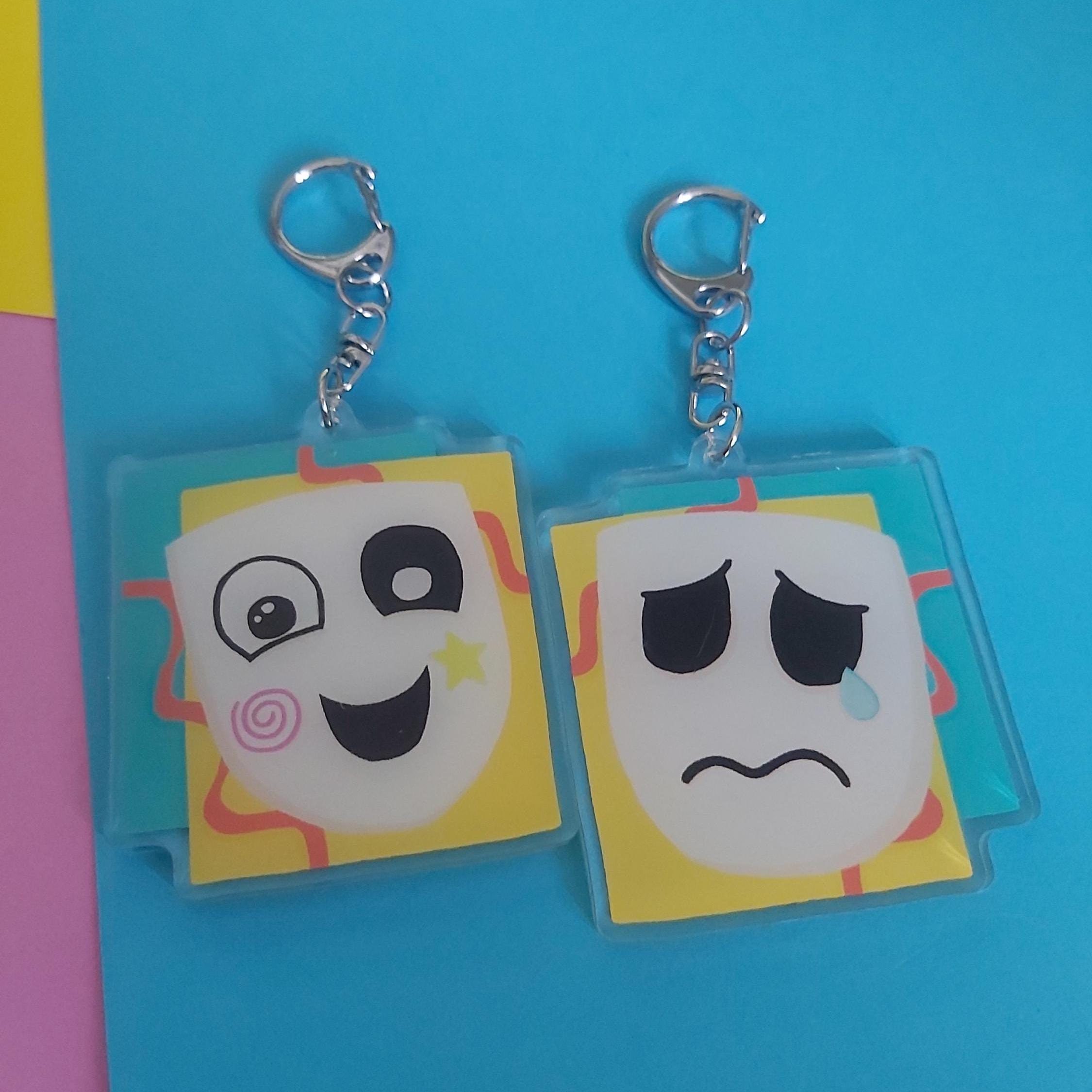 A reversible acrylic keychain with cartoon faces, one smiling and one sad, attached to silver keyrings, displayed on a bright blue background.
