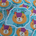A close-up of a die-cut sticker featuring a cartoon bear face with the text 'Just a little guy' on a light blue cloud-shaped background. The bear has an orange face, a purple nose, and small multicoloured ears.