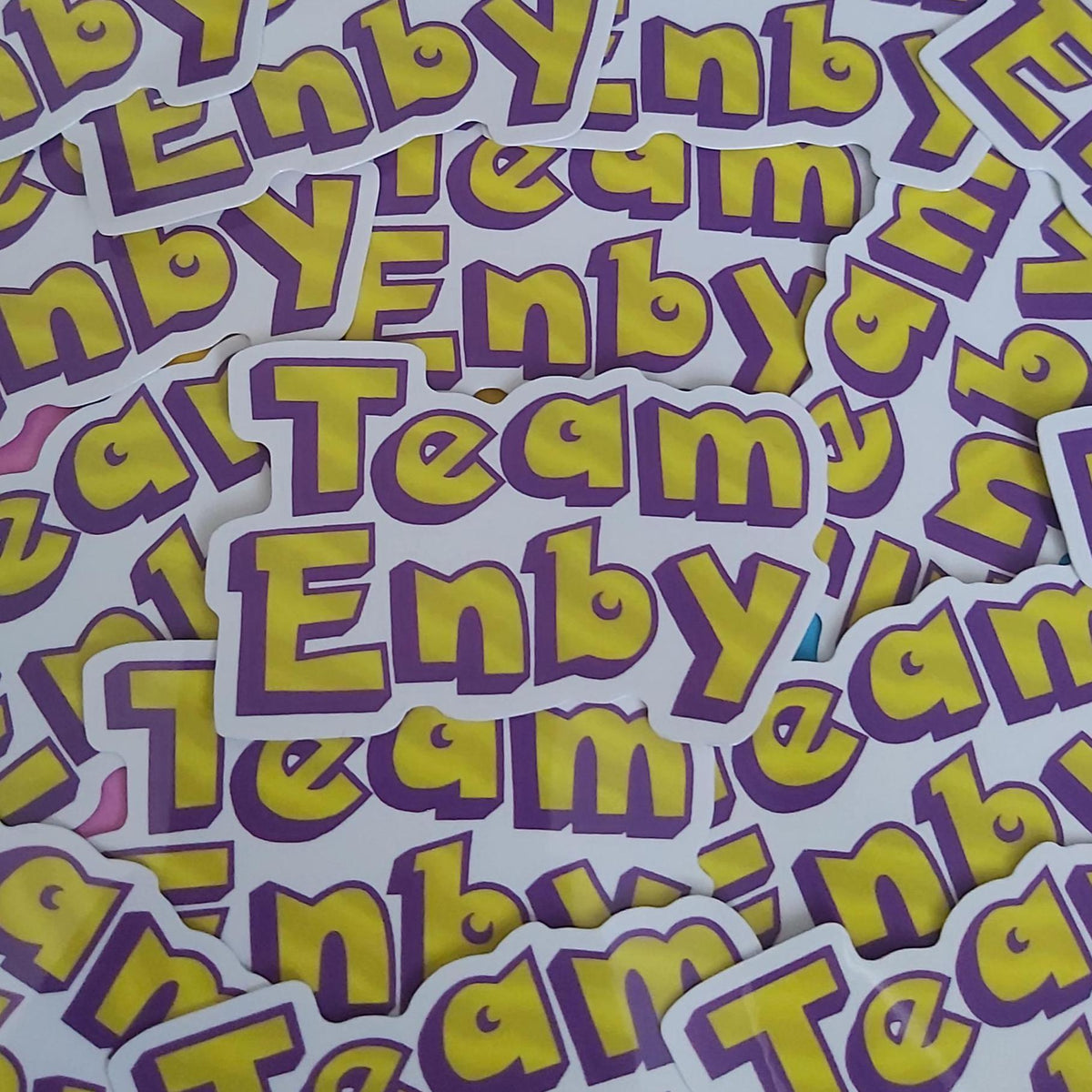 A pile of die-cut stickers featuring the text 'Team Enbya' in bold yellow letters with a purple outline on a white background.