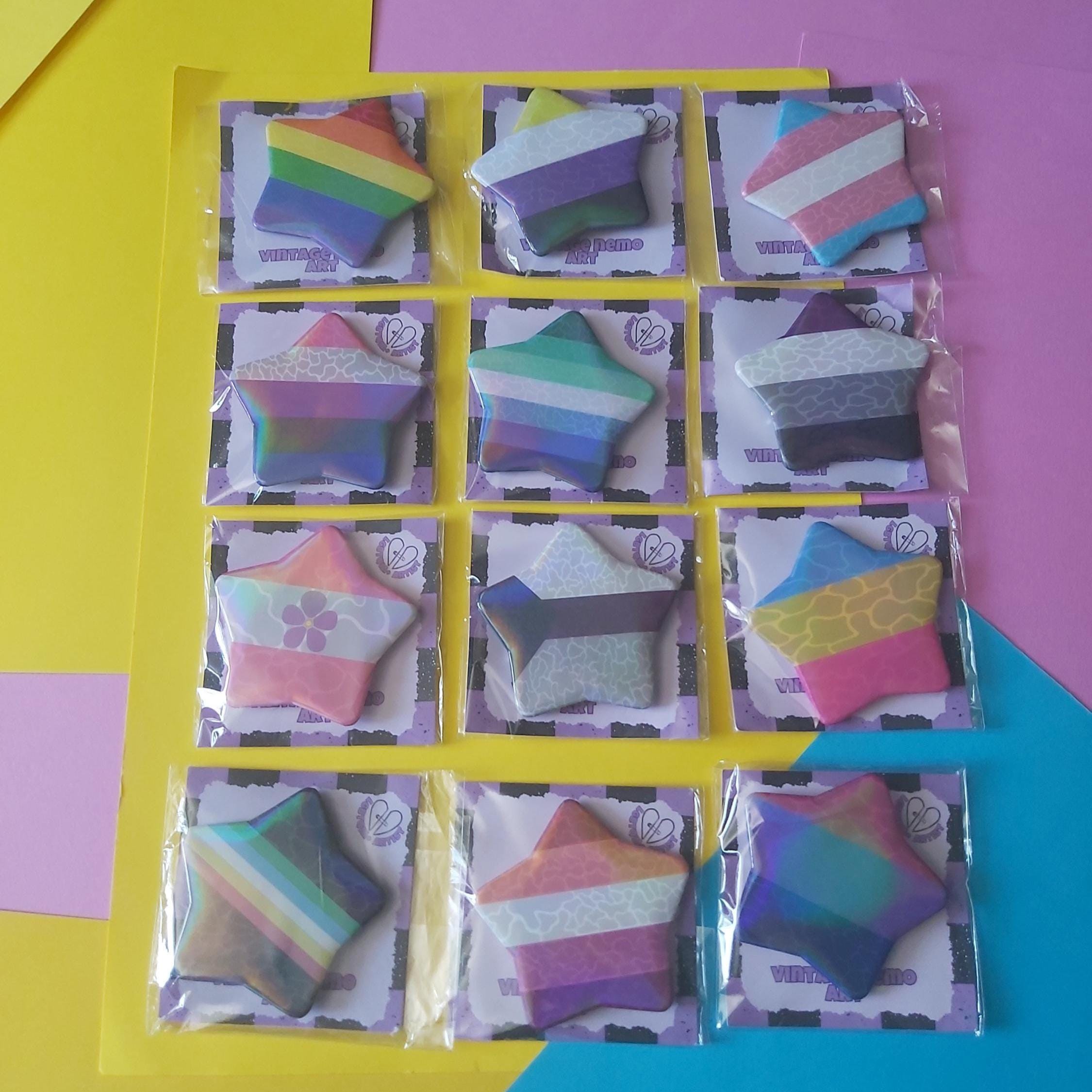Twelve star-shaped badges with various pride flags, each individually sealed in clear plastic packaging and arranged in a 3x4 grid on a yellow sheet, set against a background of yellow, pink, and blue geometric shapes.