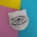 A white cat-head-shaped pin with black text reading 'The Cat Whiskers Come From Within' and a black whisker-and-heart graphic, photographed on a colourful pastel background split into pink, yellow, and blue sections.