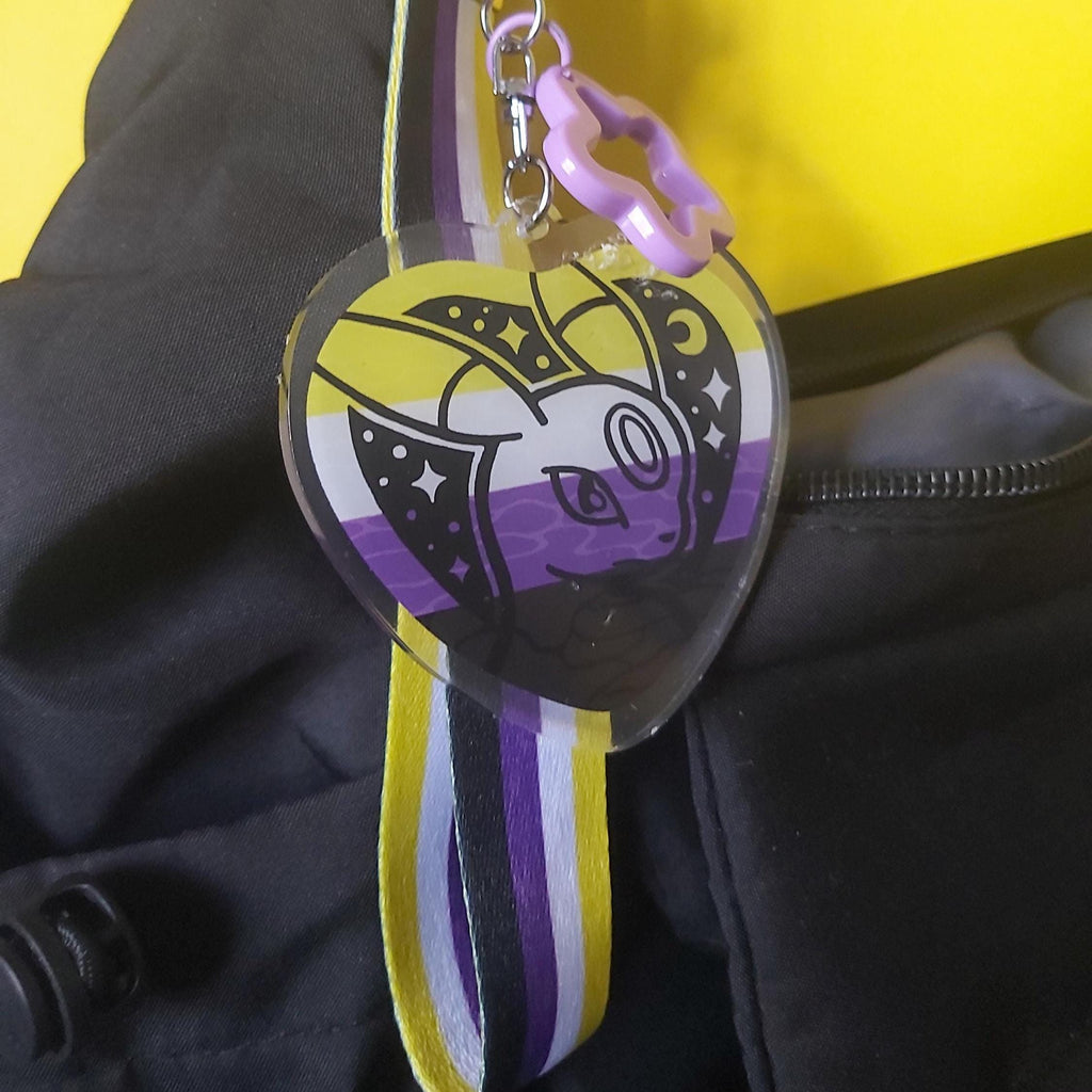 A heart-shaped acrylic keychain bag charm featuring a stylised Pokémon cartoon face in yellow, purple, black, and white, attached to a matching striped purple, yellow, black, and white strap with a purple ring clasp, shown resting on a black bag against a bright yellow background.
