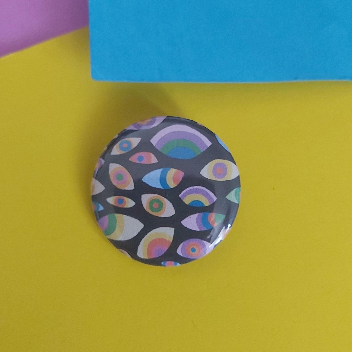 A round pinback button featuring a colourful pattern of stylised eyes and rainbows on a dark background, placed on a bright yellow surface with a blue paper corner visible at the top.