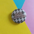 A round pin badge with a black and white checkered pattern and the text 'OFF TO COMMIT TOMFOOLERY' in red, placed on a background divided diagonally into yellow, blue, and pink sections.