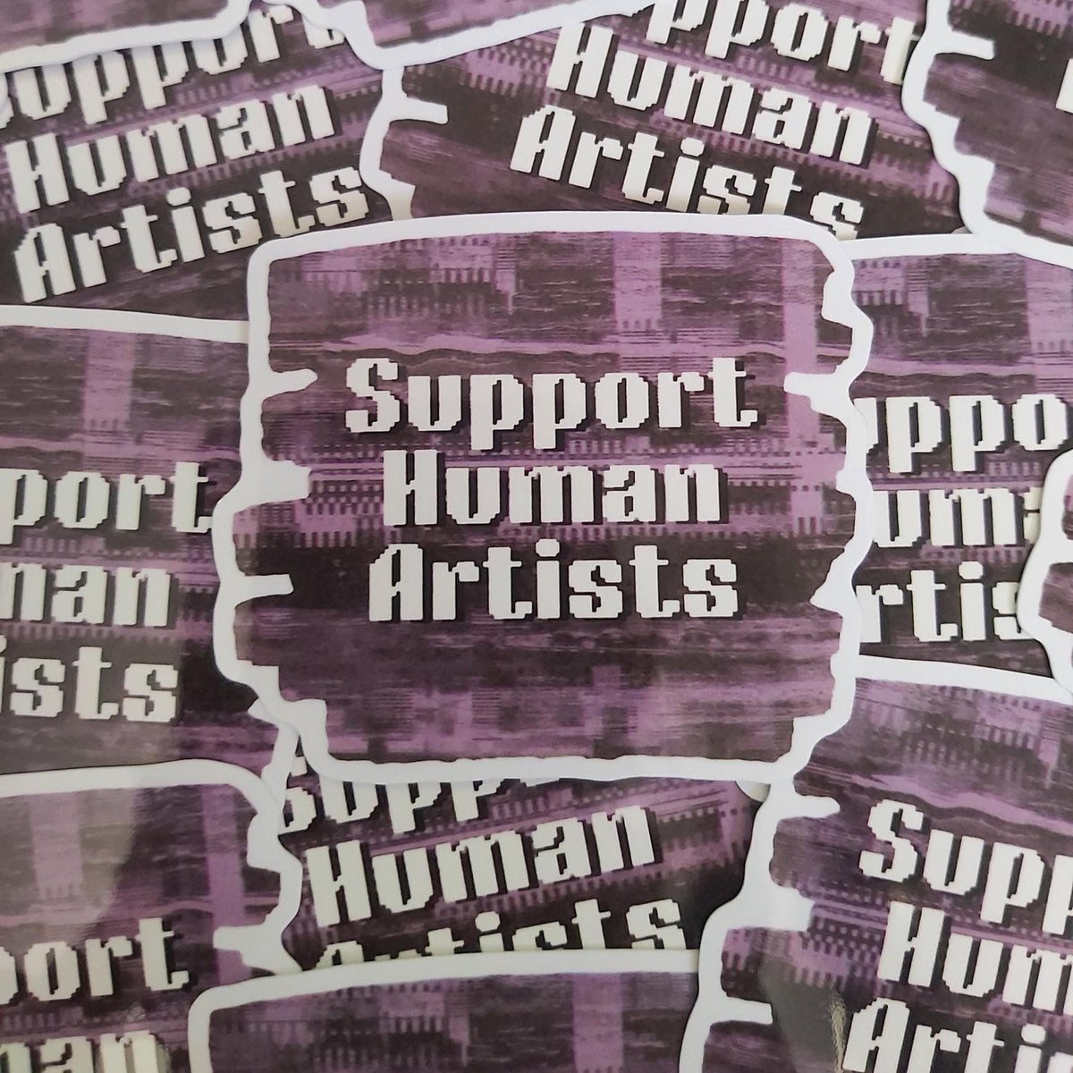A close-up of multiple die-cut stickers with the phrase 'Support Human Artists' in white text over a textured purple and black background, arranged with overlapping matching stickers.
