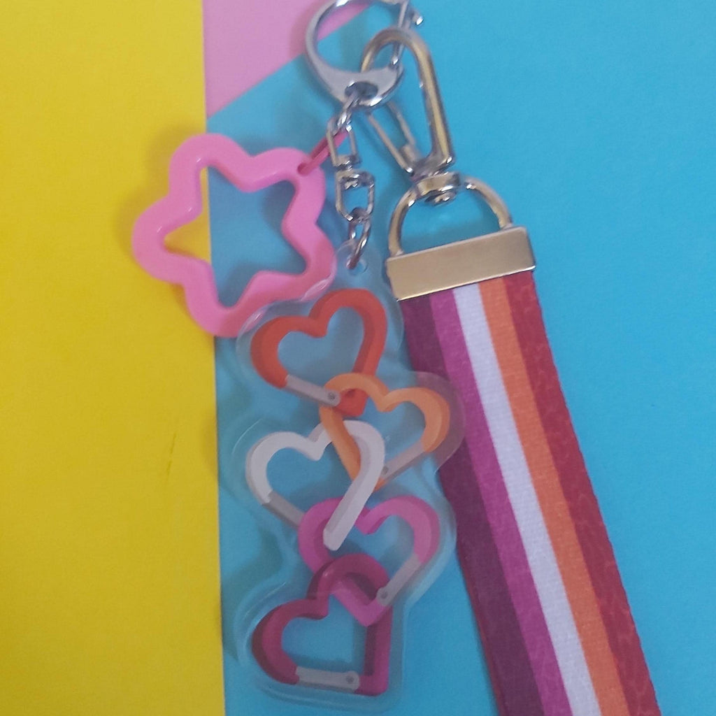 A colourful keychain with a pink star-shaped charm, an acrylic charm of interlocking heart carabiners in pink, orange, white, and magenta, a silver lobster clasp, and a striped fabric ribbon in pink, orange, white, and magenta.