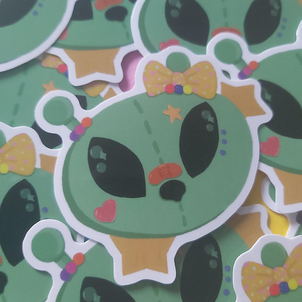 A pile of die-cut stickers featuring a cute green alien character with a yellow polka-dot bow tie, black eyes, and small colourful accents.