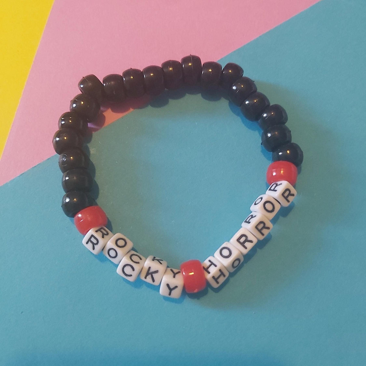 A beaded stretch bracelet featuring glossy black round beads, red accent beads, and white letter cube beads spelling out 'ROCKY HORROR', displayed on a colorful background of pink, yellow, and blue paper.