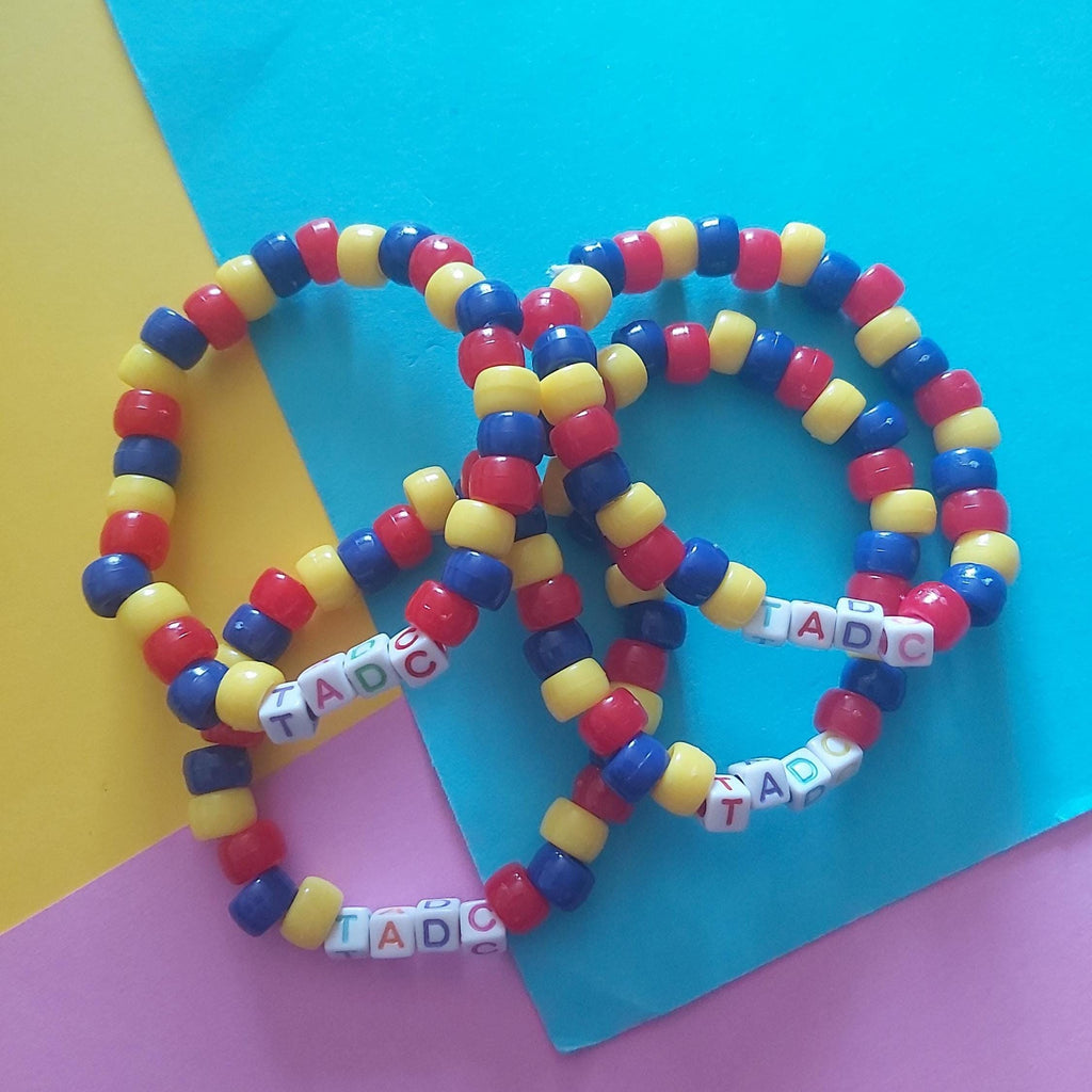 Four beaded stretch bracelets arranged on a colourful background. The bracelets are made of alternating red, yellow, and blue round beads, with white letter beads spelling 'TADC'.