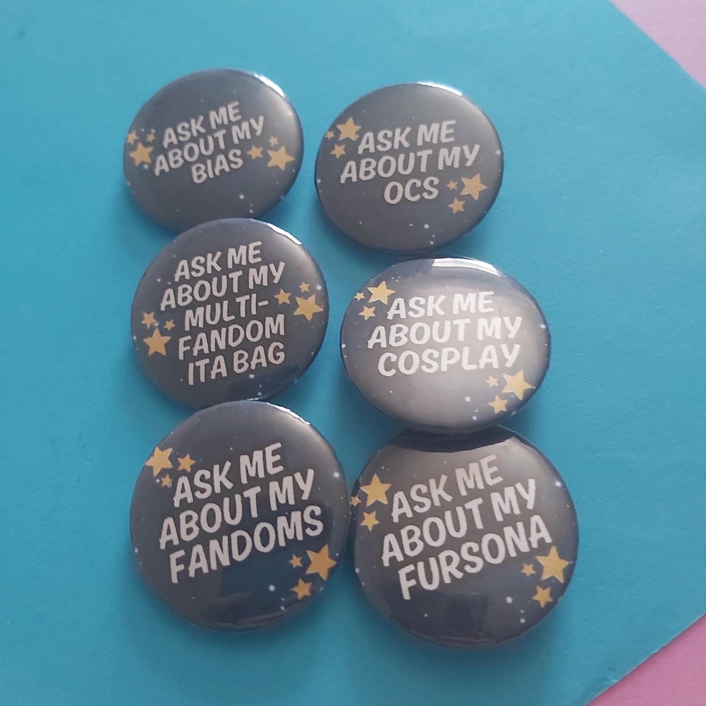 Six round pin-back buttons with dark grey backgrounds and white text reading phrases like 'ASK ME ABOUT MY BIAS', 'ASK ME ABOUT MY OCS', and 'ASK ME ABOUT MY FANDOMS', decorated with small gold stars, arranged on a bright blue background.