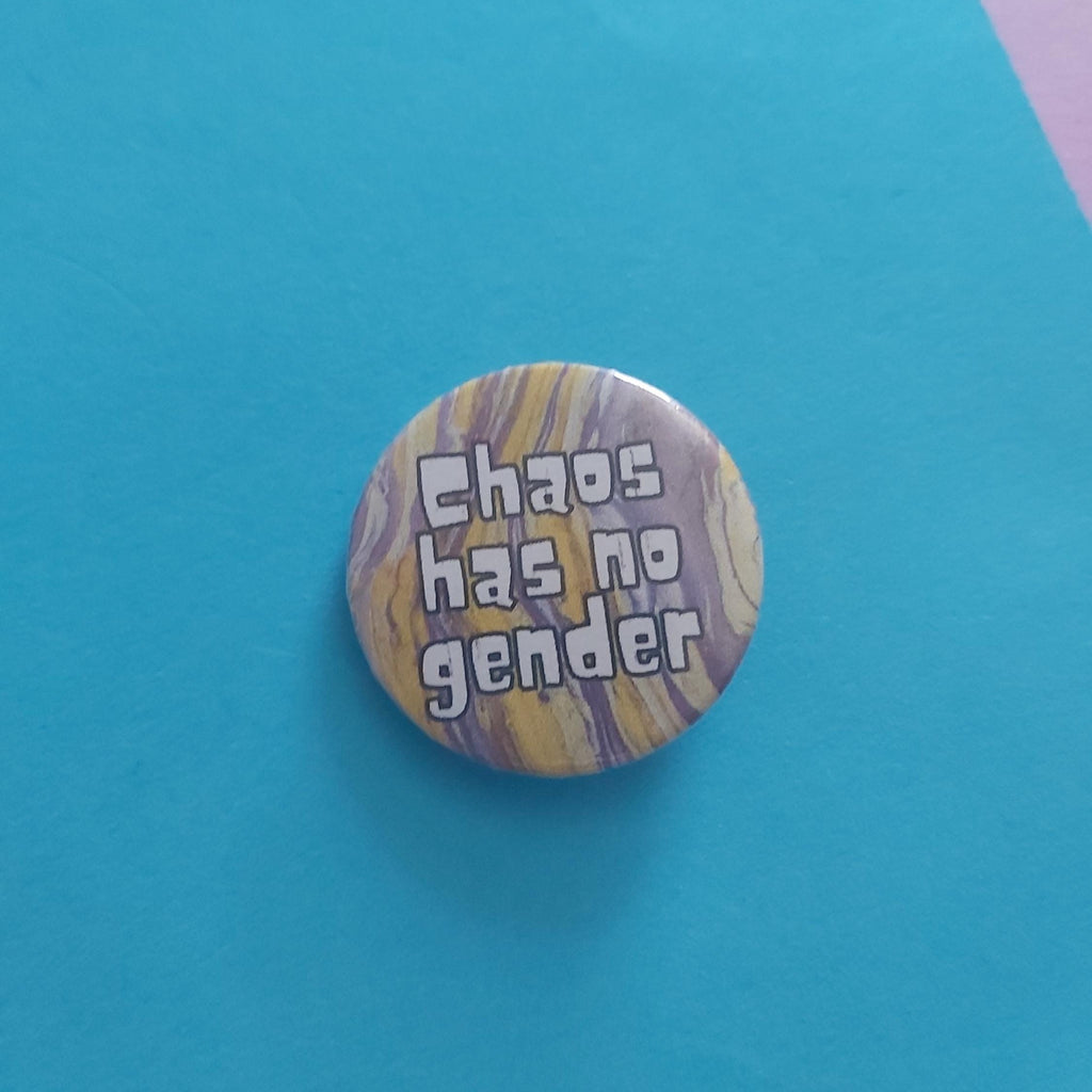 A round pinback button badge featuring a yellow and purple marbled pattern with the text 'Chaos has no gender' in white letters, displayed on a bright blue background.