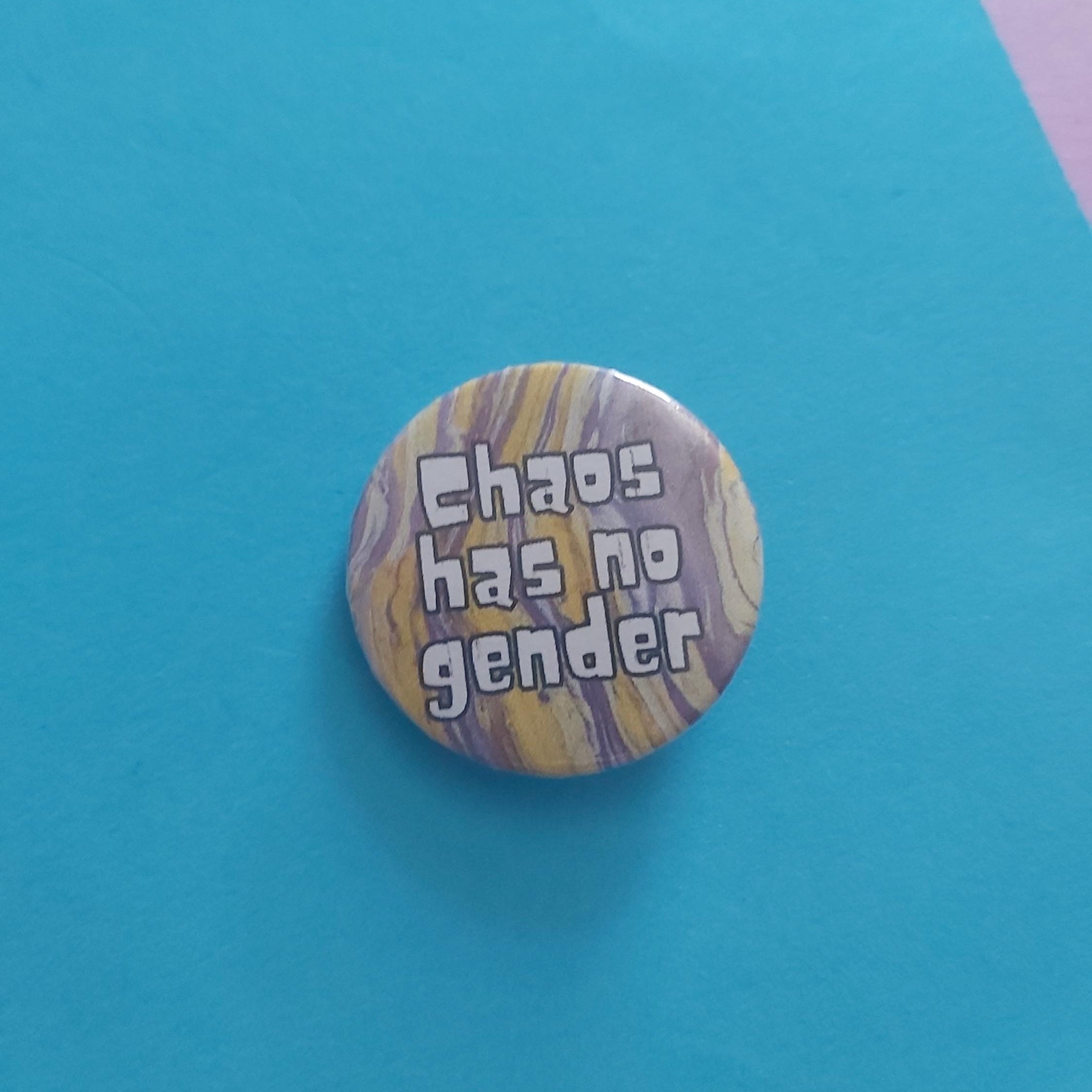 A round pinback button badge featuring a yellow and purple marbled pattern with the text 'Chaos has no gender' in white letters, displayed on a bright blue background.