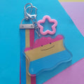 A striped keychain charm with a pink star-shaped ring and silver clasp, photographed on a colourful geometric paper background.