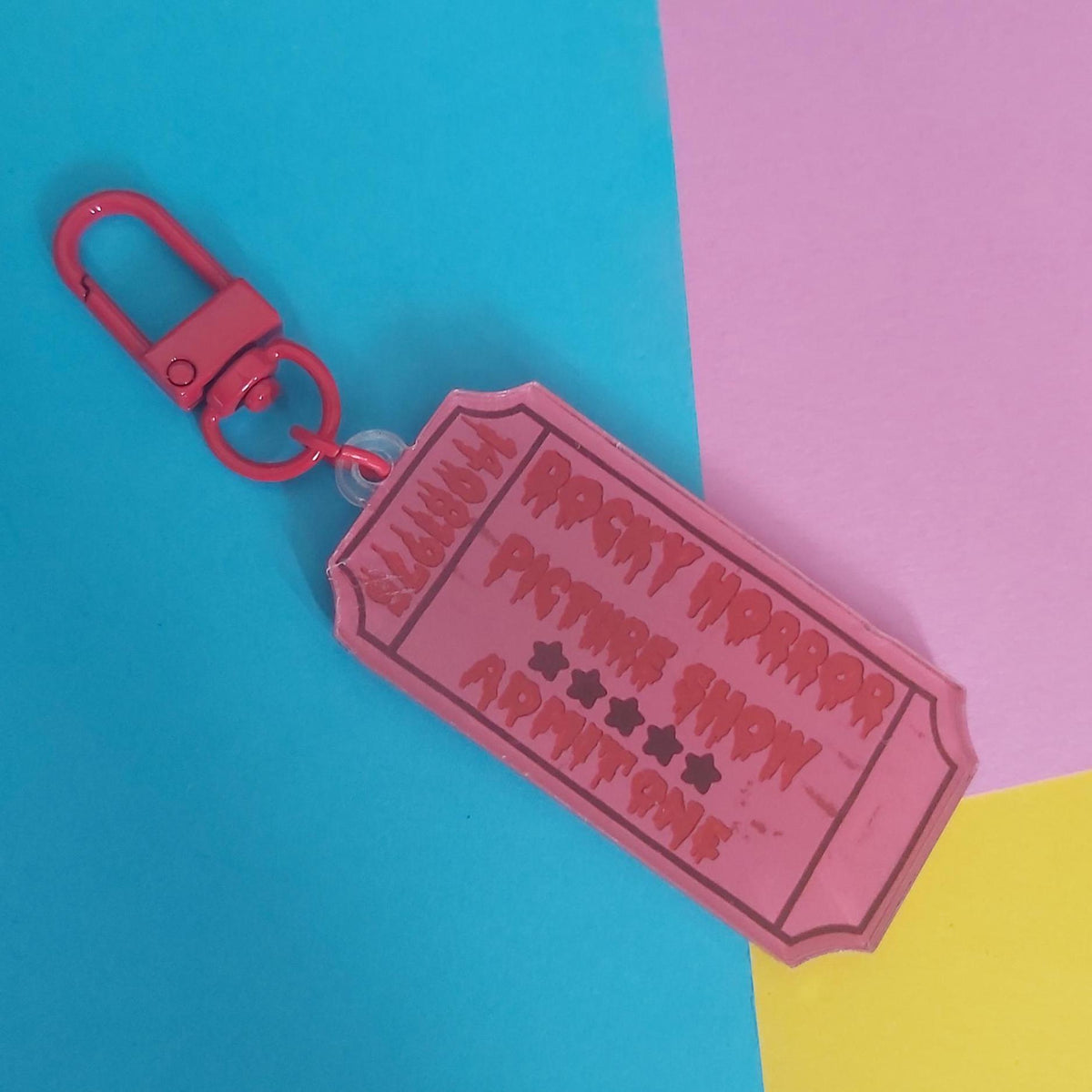 A pink ticket-shaped keychain with a red carabiner clip, featuring the text 'ROCKY HORROR PICTURE SHOW ADMIT ONE' on a split blue, pink, and yellow background.
