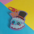A colourful acrylic keychain shaped like Caine from The Amazing Digital Circus wearing a black top hat, attached to a red clasp, photographed on a bright diagonally split yellow, pink, and blue background.