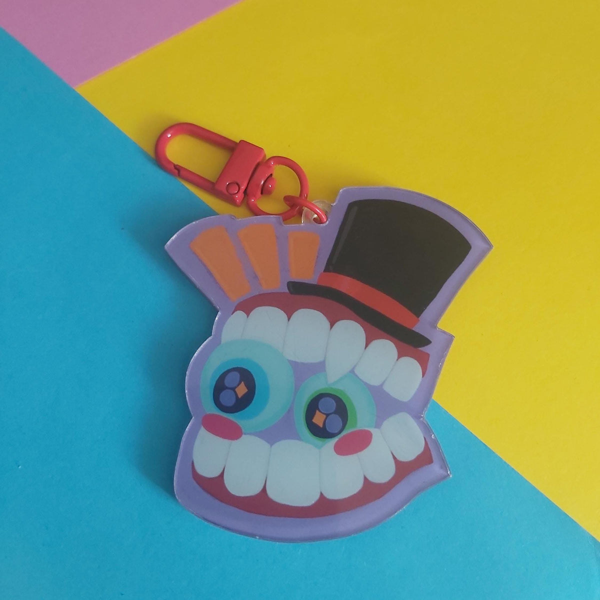 A colourful acrylic keychain shaped like Caine from The Amazing Digital Circus wearing a black top hat, attached to a red clasp, photographed on a bright diagonally split yellow, pink, and blue background.