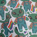 A close-up of multiple die-cut stickers featuring a green alien character wearing rainbow-striped overalls and holding a small rainbow flag, arranged on a white background.
