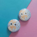Two round pinback buttons featuring a cartoon character with large eyes, displayed on a diagonally split teal and pink background.