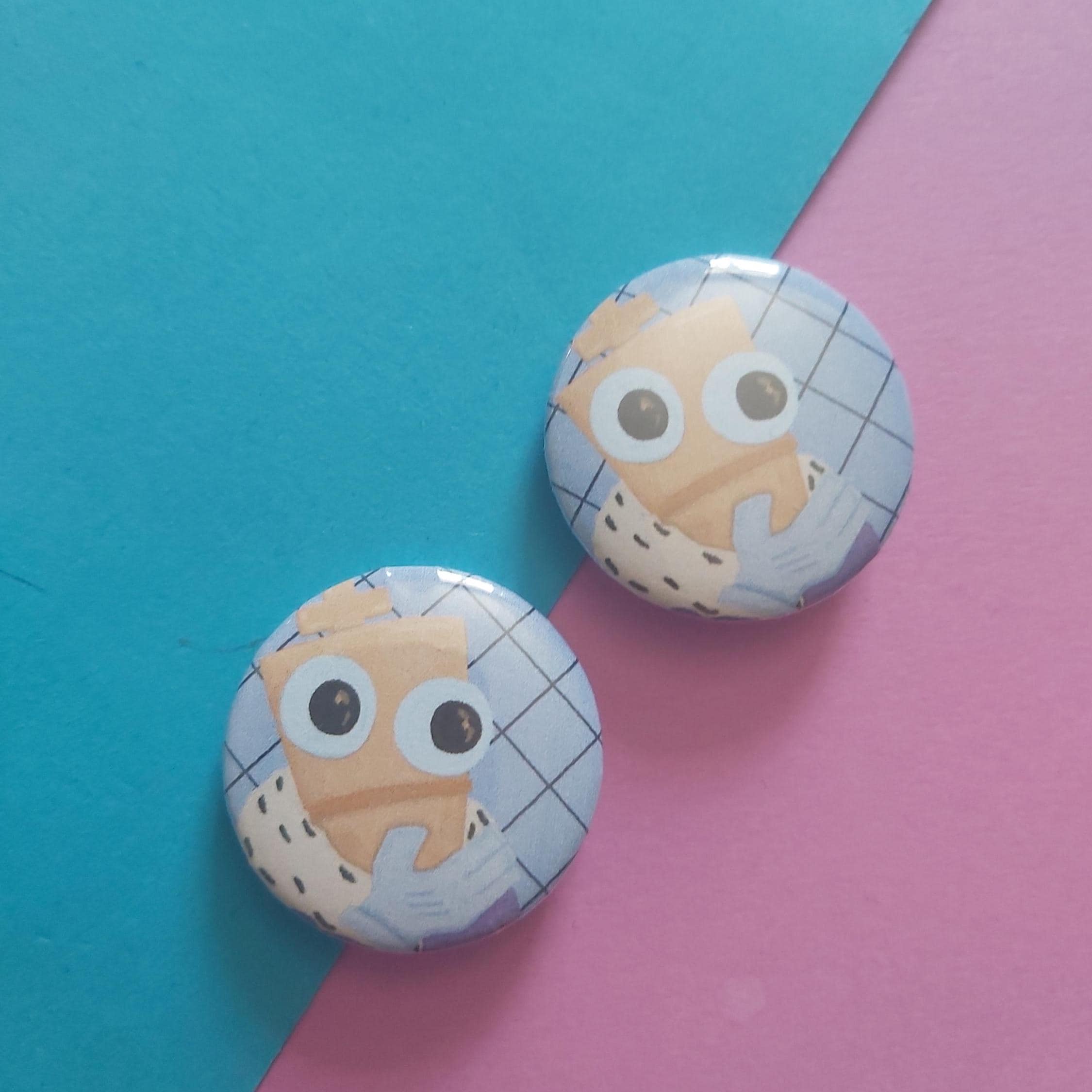 Two round pinback buttons featuring a cartoon character with large eyes, displayed on a diagonally split teal and pink background.