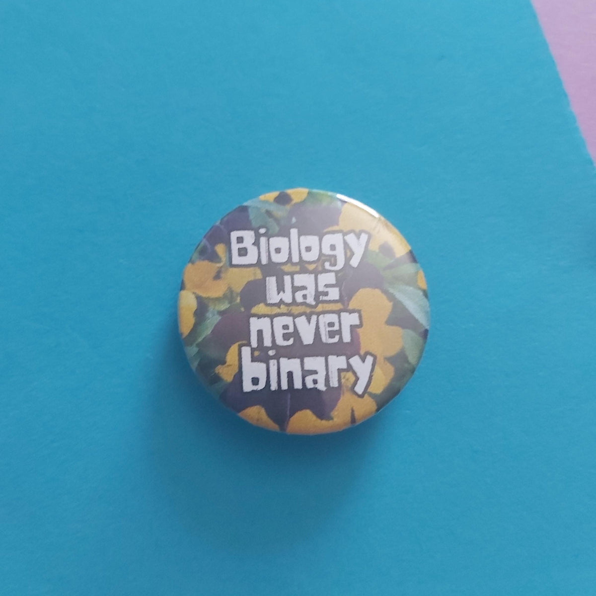 A round pinback button with the phrase 'Biology was never binary' in white text over a yellow, orange, and dark gray camouflage pattern, displayed on a bright blue background.