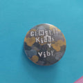 A round pinback button with a floral camouflage pattern in yellow, grey, and black, featuring the text 'GENDER KILLS MY VIBE' in white. The pin is displayed on a bright blue background with a corner of pink paper visible in the bottom right.