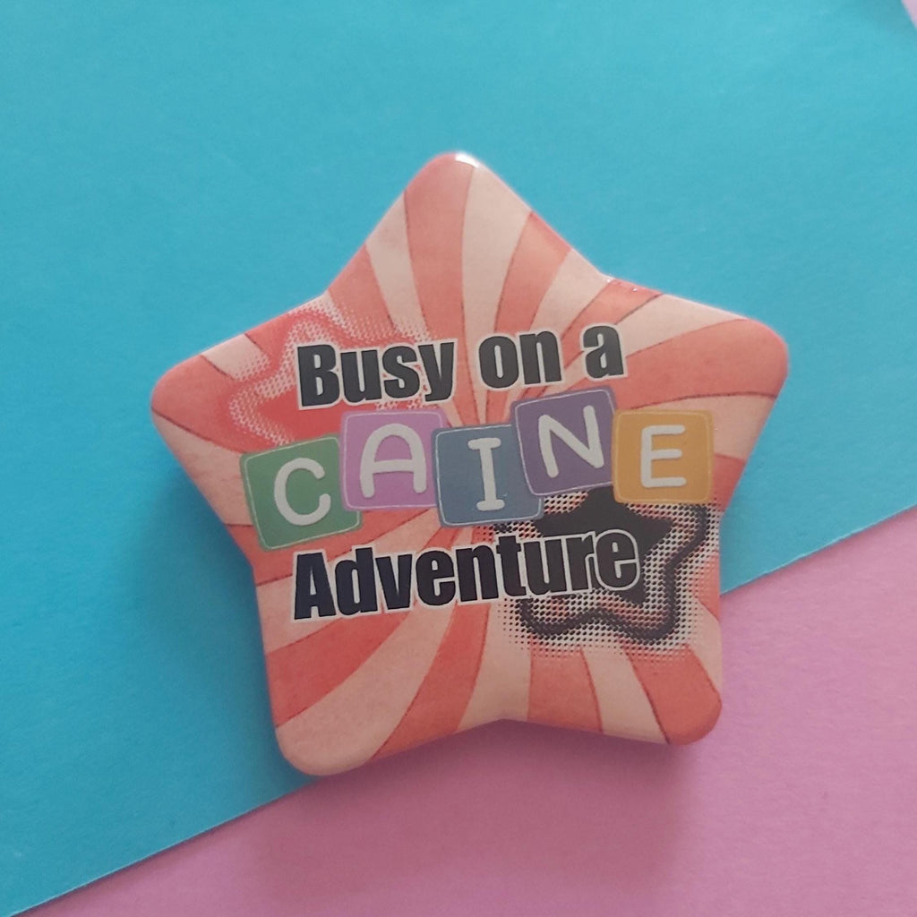 A star-shaped pin badge with a red and white sunburst pattern, featuring the text 'Busy on a Caine Adventure' in black and colourful block letters