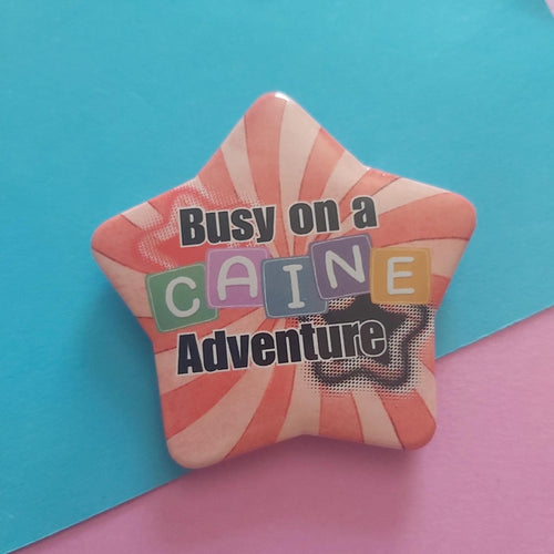 A star-shaped pin badge with a red and white sunburst pattern, featuring the text 'Busy on a Caine Adventure' in black and colourful block letters