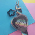 A heart-shaped acrylic keychain featuring a Pokémon character, attached to a striped strap and a black star-shaped charm, displayed on a colourful background of blue, pink, and yellow paper.