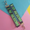 Two black fabric keychains with silver-tone clasp hardware, featuring the text 'I <3 ALIENS' in bright green letters, displayed on a colourful pink, blue, and yellow background.