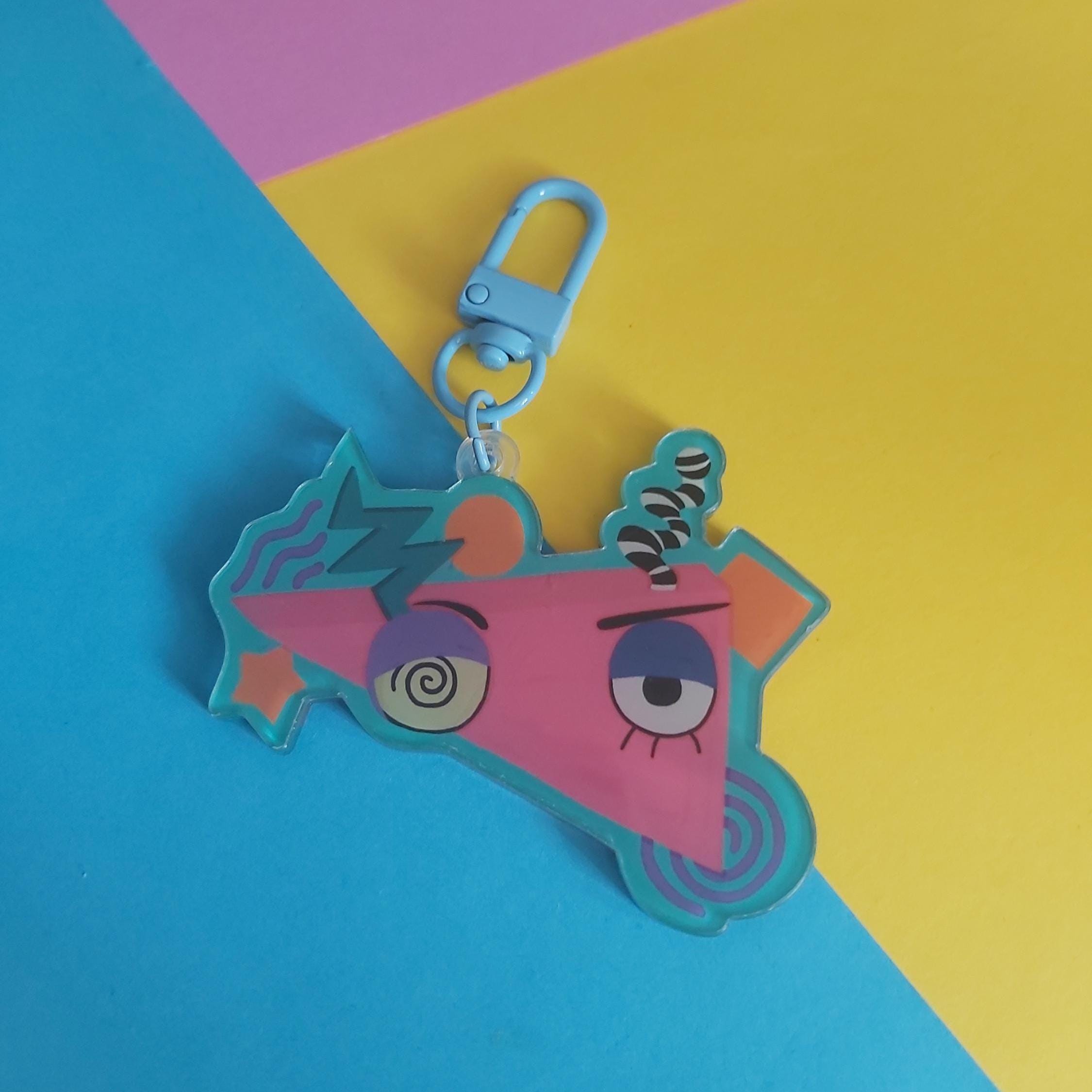A colourful acrylic keychain charm featuring a stylised cartoon face with one spiralling eye and one wide eye, attached to a light blue lobster clasp. The charm is displayed on a diagonally split yellow and blue background.