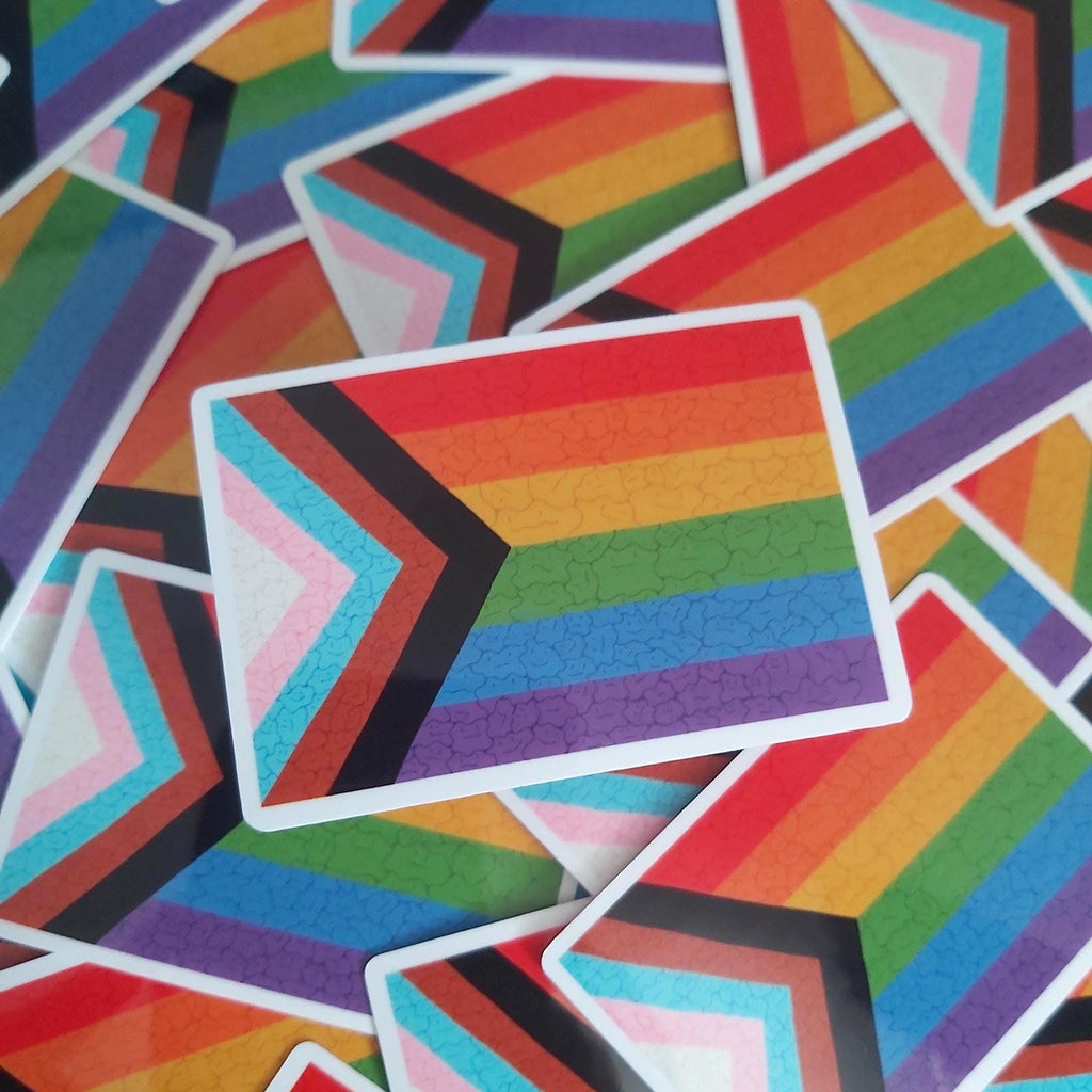 A pile of rectangular stickers featuring the Progress Pride flag design with rainbow stripes and chevron overlays, photographed closely together.