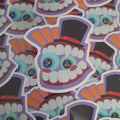 A close-up pile of colourful die-cut stickers featuring a cartoon character called Caine's face with large white teeth, blue and green eyes, orange and purple accents, and black top hats.