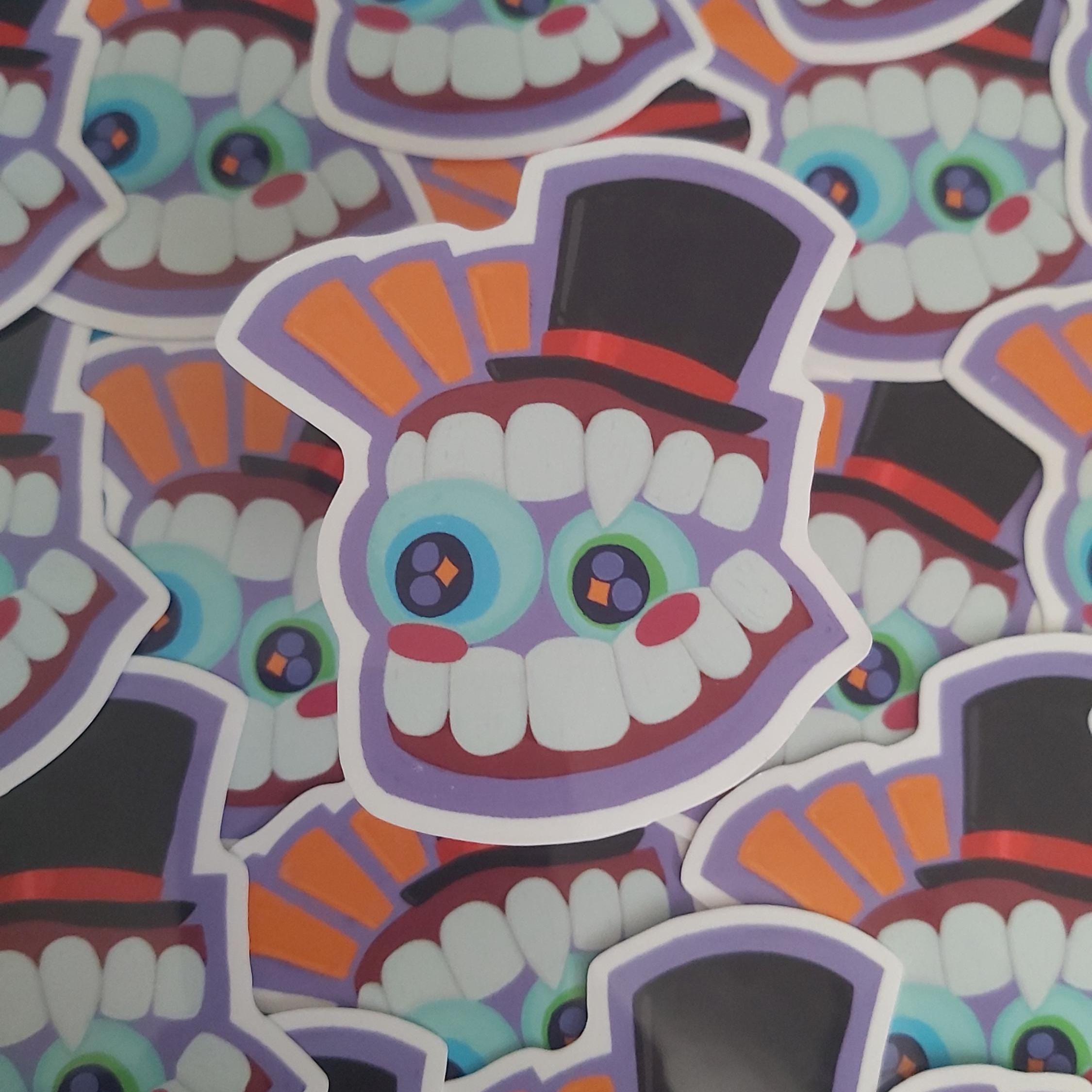 A close-up pile of colourful die-cut stickers featuring a cartoon character called Caine's face with large white teeth, blue and green eyes, orange and purple accents, and black top hats.
