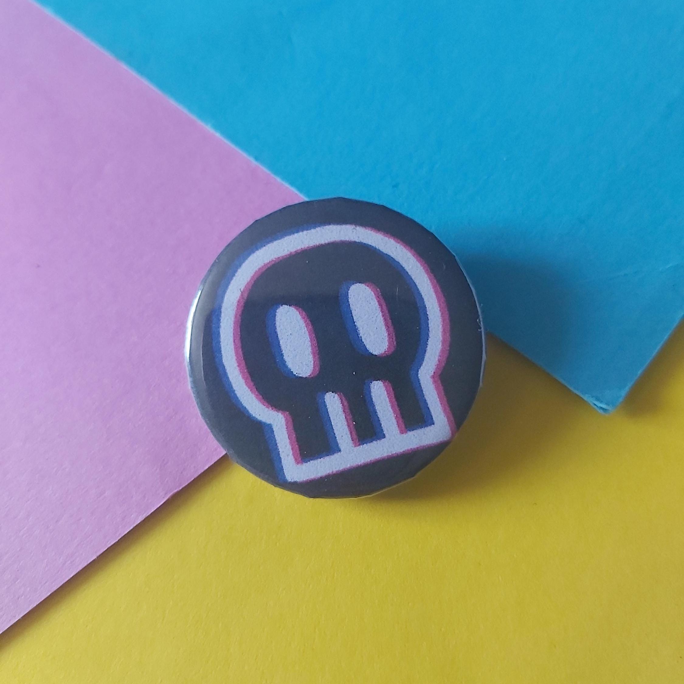 A round pin badge featuring a stylised white skull with pink and blue glitch-style outlines on a dark black background, set against a colourful background of pink, blue, and yellow paper.