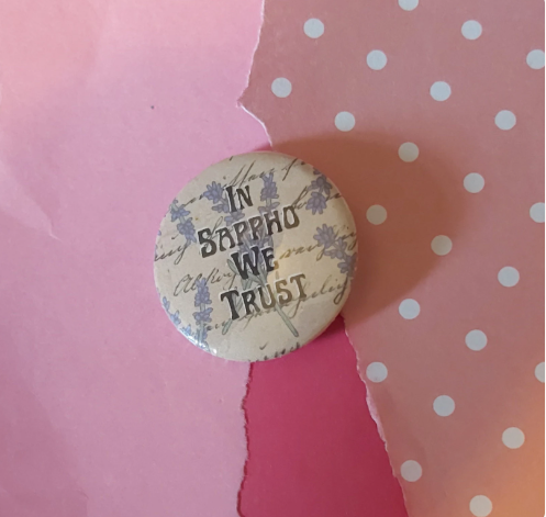 A round pin-back button badge with a vintage-style cream background, light purple floral pattern, and cursive script, featuring the text 'IN SAPPHO WE TRUST' in dark lettering. The button is photographed on a styled background of torn pink paper and pink polka-dot paper.