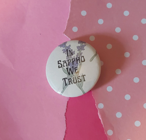 A round pin-back button badge featuring the text 'IN SAPPHO WE TRUST' in black lettering over a lavender floral illustration, placed on a pink background with torn paper and white polka dots.